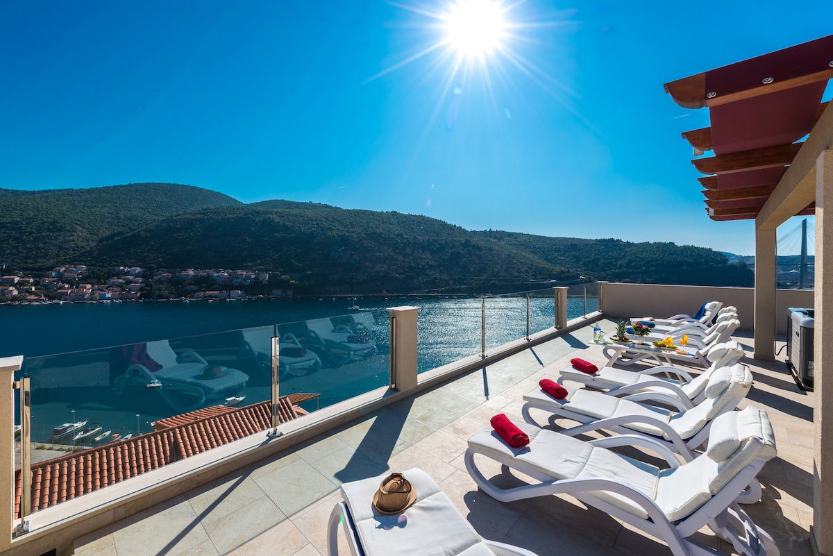 A spacious shared terrace is equipped with sun loungers, facing a stunning view of the sea and mountains. The clear sky is brightened by abundant sunlight, enhancing the relaxing atmosphere. Glass railings provide unobstructed views of the surrounding landscape.