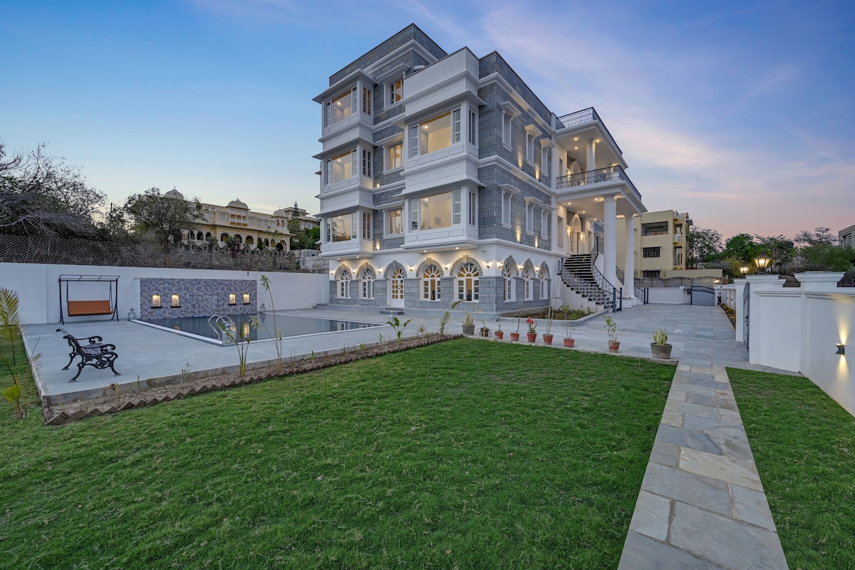 The exterior showcases a grand three-story building with layered balconies and arched windows. A well-maintained green lawn surrounds the swimming pool, complemented by landscaped pathways and seating areas, all set against a serene sky transitioning to twilight.