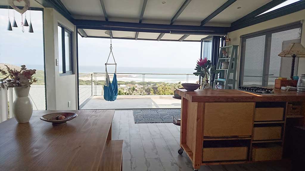 An open-concept living space features a large sliding glass door that connects to a panoramic view of the ocean. A comfortable eating area is complemented by a kitchen with modern amenities. A hanging swing chair adds a cozy touch to the serene atmosphere.