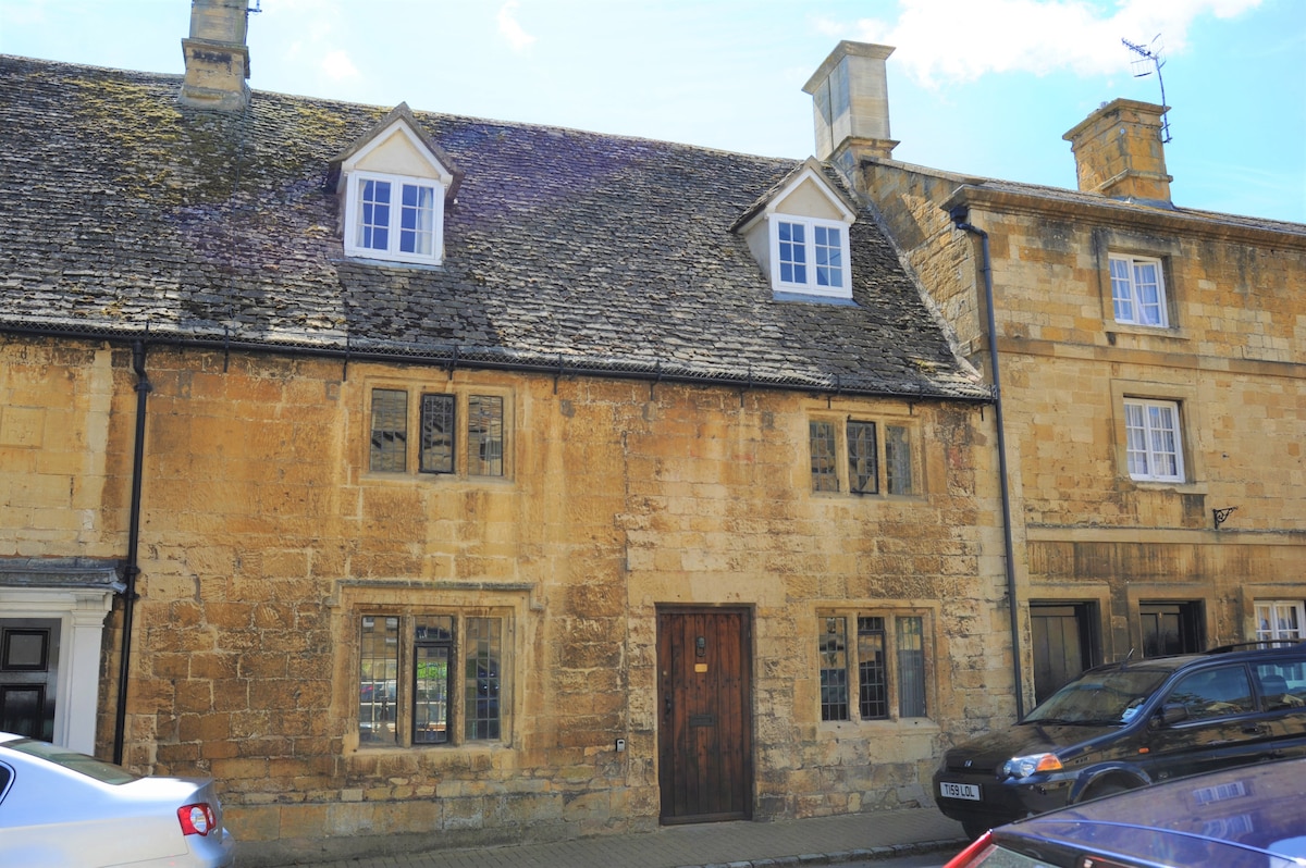 The front exterior of a Cotswold stone house is depicted, featuring a traditional design with stone walls and a slate roof. The structure includes multiple windows with white frames and wooden doors, nestled among neighboring buildings and parked cars along the street.