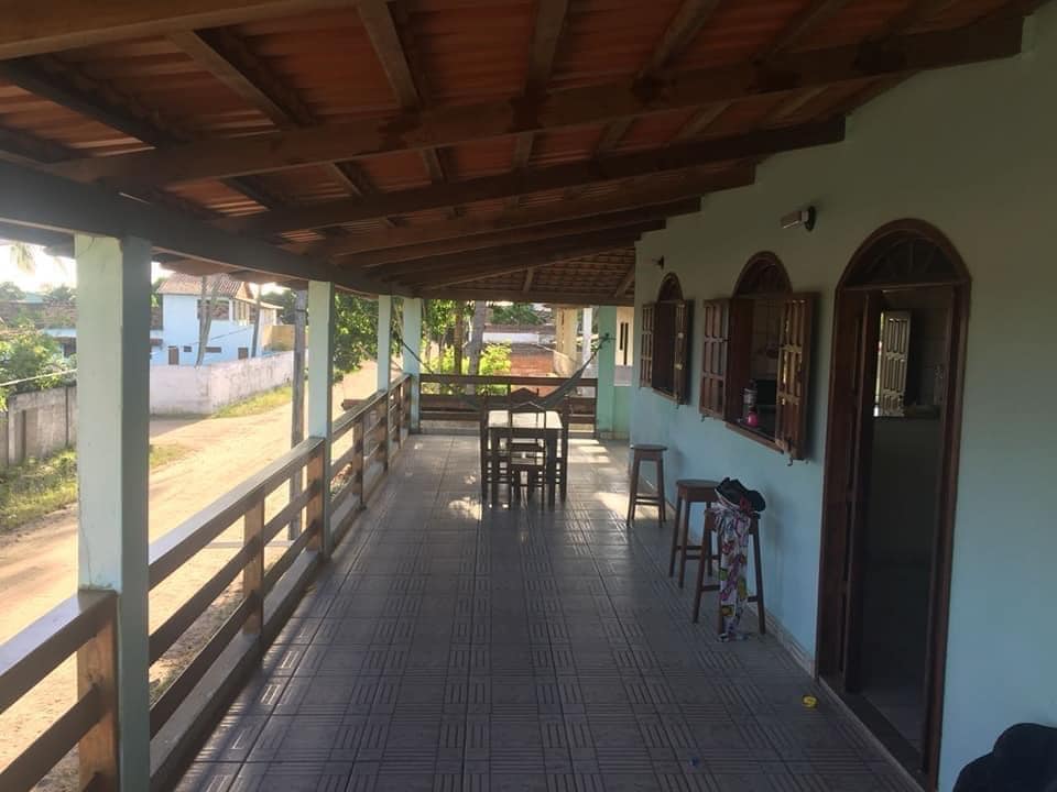 A covered balcony extends along the house, featuring a wooden railing and a table with four chairs. The tiled floor reflects ample natural light, while the surrounding greenery and road visibility create a serene outdoor space. Access to the interior is visible through open doorways.