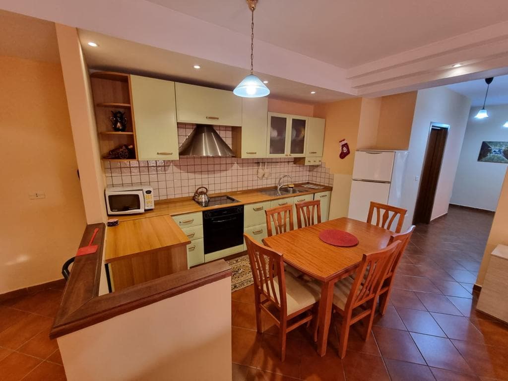 The kitchen area features light cabinetry with modern appliances, including a stovetop and microwave. A wooden dining table with six chairs is positioned nearby. Soft lighting creates a warm ambiance, and tiles cover the floor for practicality and ease of cleaning.