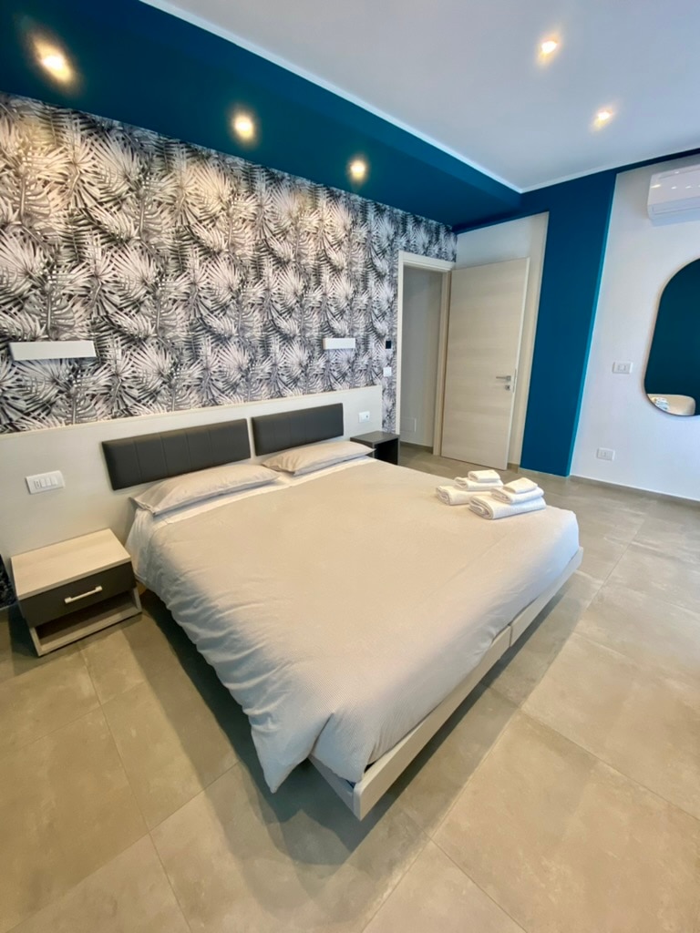 A modern bedroom features a spacious bed with light bedding and minimalistic decor. A patterned accent wall with palm leaves provides contrast. Nightstands frame the bed, and ambient lighting enhances the room. A mirror and a door lead to the bathroom, with a cool color scheme throughout.