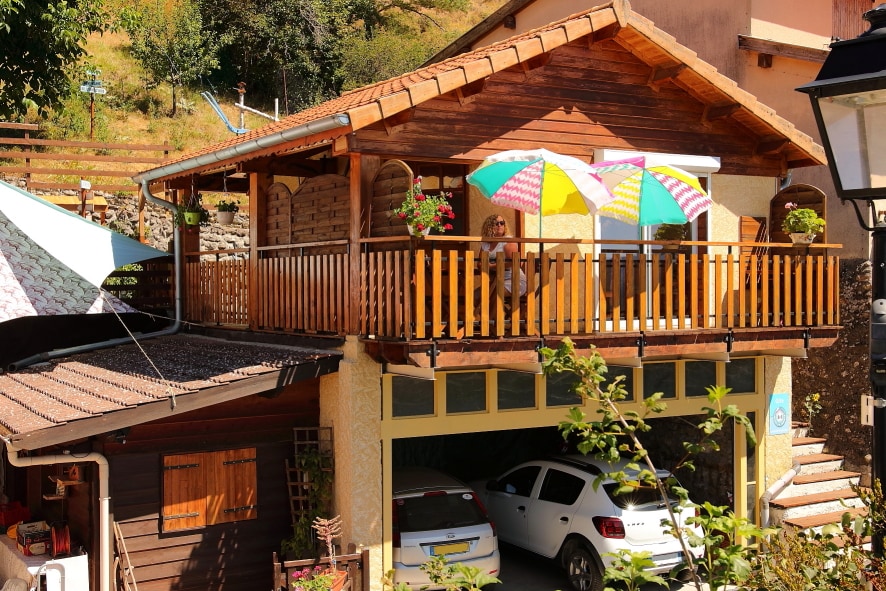 A wooden chalet with a terrace showcases a warm, inviting entrance, featuring colorful umbrellas and flower pots. Below, a garage accommodates two vehicles, with lush greenery and a small pathway visible, enhancing the chalet's peaceful mountain setting.