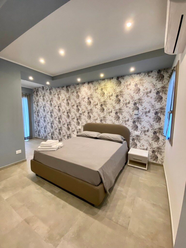 A modern bedroom features a spacious bed dressed in neutral linens, complemented by a bedside table. A floral accent wall adds subtle character. Natural light filters through a window, with a view of the private terrace, enhancing the room's airy feel.
