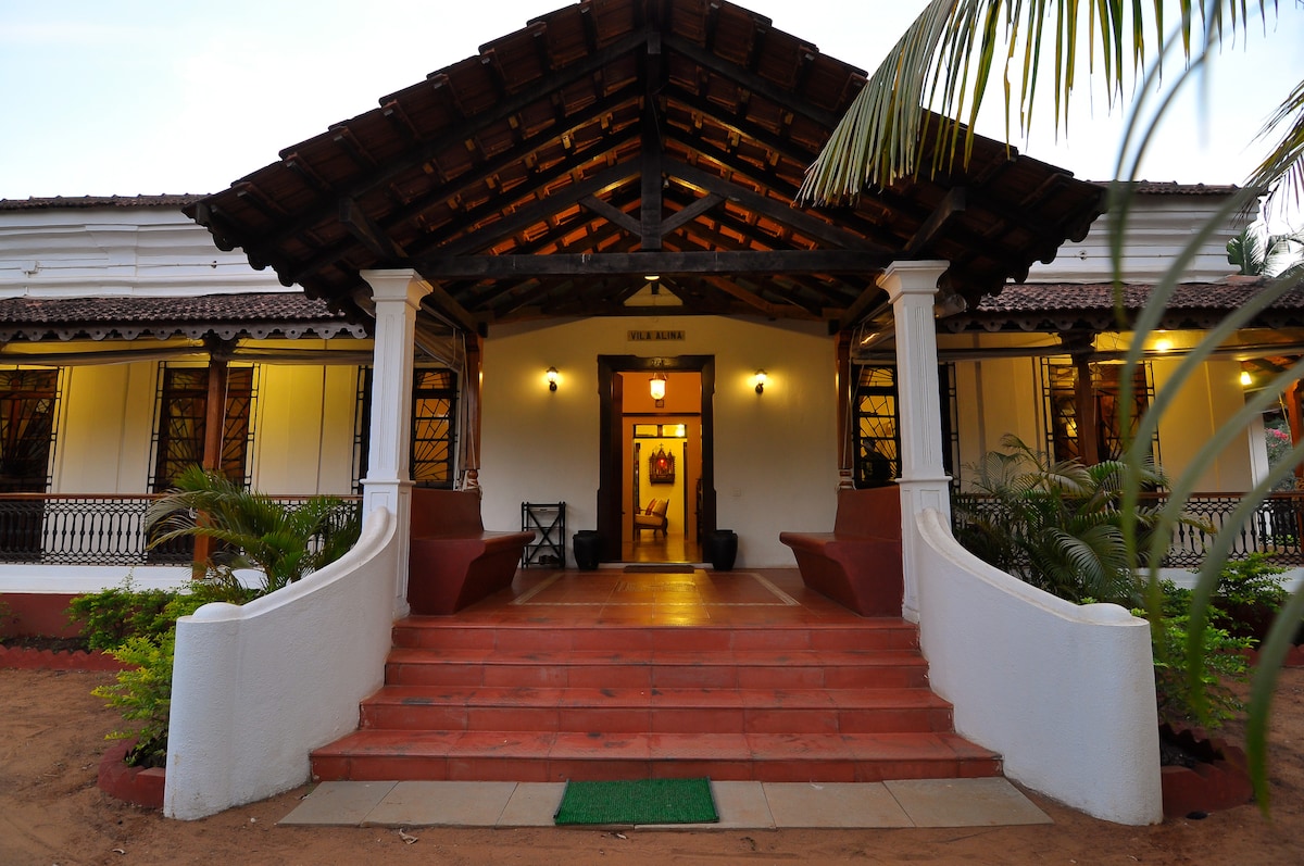 The entrance of the villa is framed by a wooden roof with intricate beams, welcoming guests with a spacious pathway. Surrounding greenery and palm trees enhance the setting, while traditional seating options invite relaxation near the entrance.