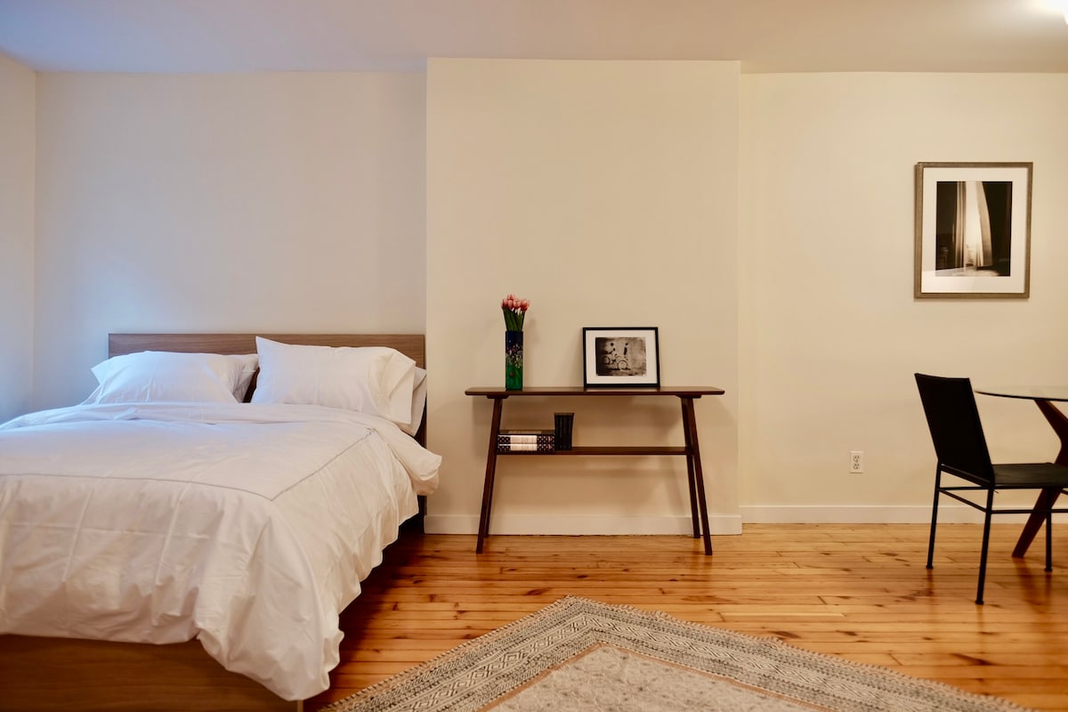 A queen-sized bed with a white comforter is positioned against a light wall. A dark wooden table stands nearby, featuring a framed photo and a small vase of flowers. A black chair is situated next to the table, complementing the warm wooden flooring.