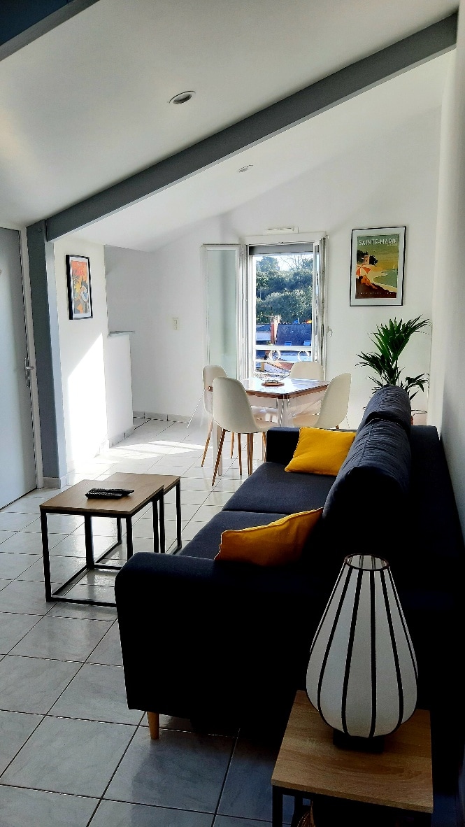 A bright living space is displayed, featuring a dark sofa with yellow accent pillows. A small coffee table is positioned in front, while a dining table with white chairs can be seen through the open balcony doors, leading to a view of greenery and outdoor seating.