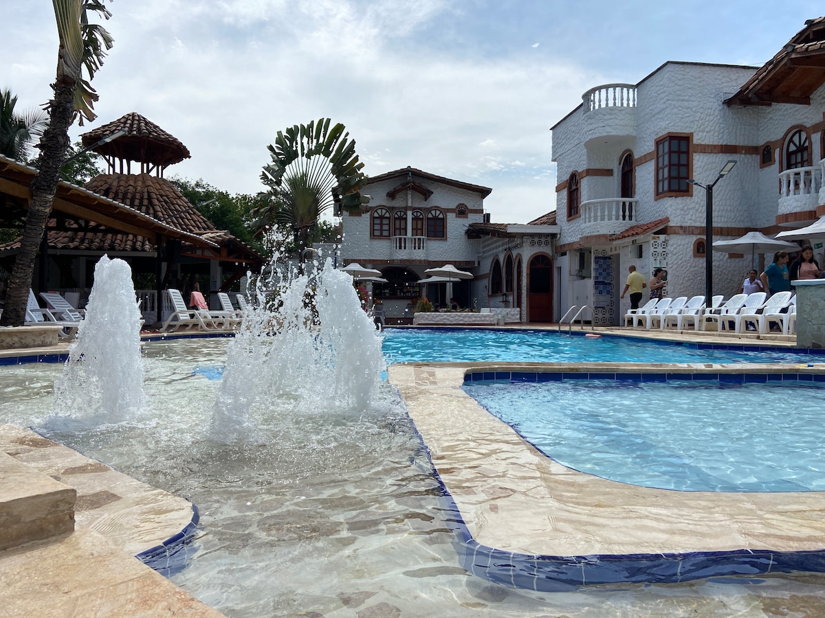 A well-maintained outdoor pool area features a splash fountain, surrounded by sun loungers. Brick and white-washed structures approximate a colonial style, with palm trees framing the scene. The inviting pool reflects the cloudy sky above.