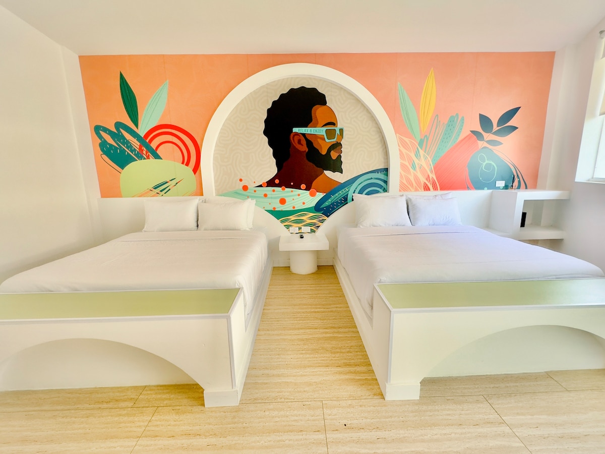 A brightly colored guest room features two spacious beds, each dressed in crisp white linens. A large mural depicting a man with sunglasses adorns the wall behind the beds, complemented by vibrant, abstract foliage designs.