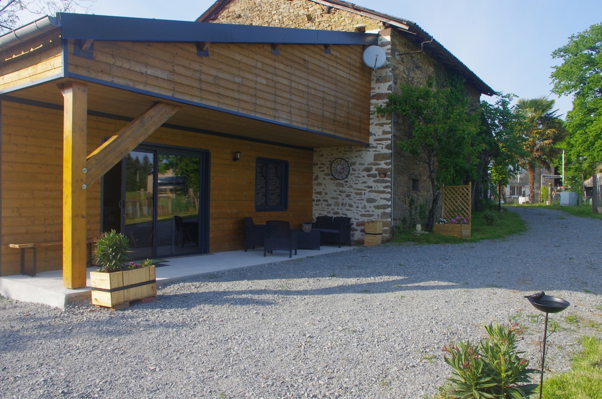 The exterior features a combination of modern wooden elements and rustic stone, creating a welcoming entrance. A gravel pathway leads from the front to the entrance area, covered by a spacious porch with seating. Greenery surrounds the building, enhancing the natural ambiance.