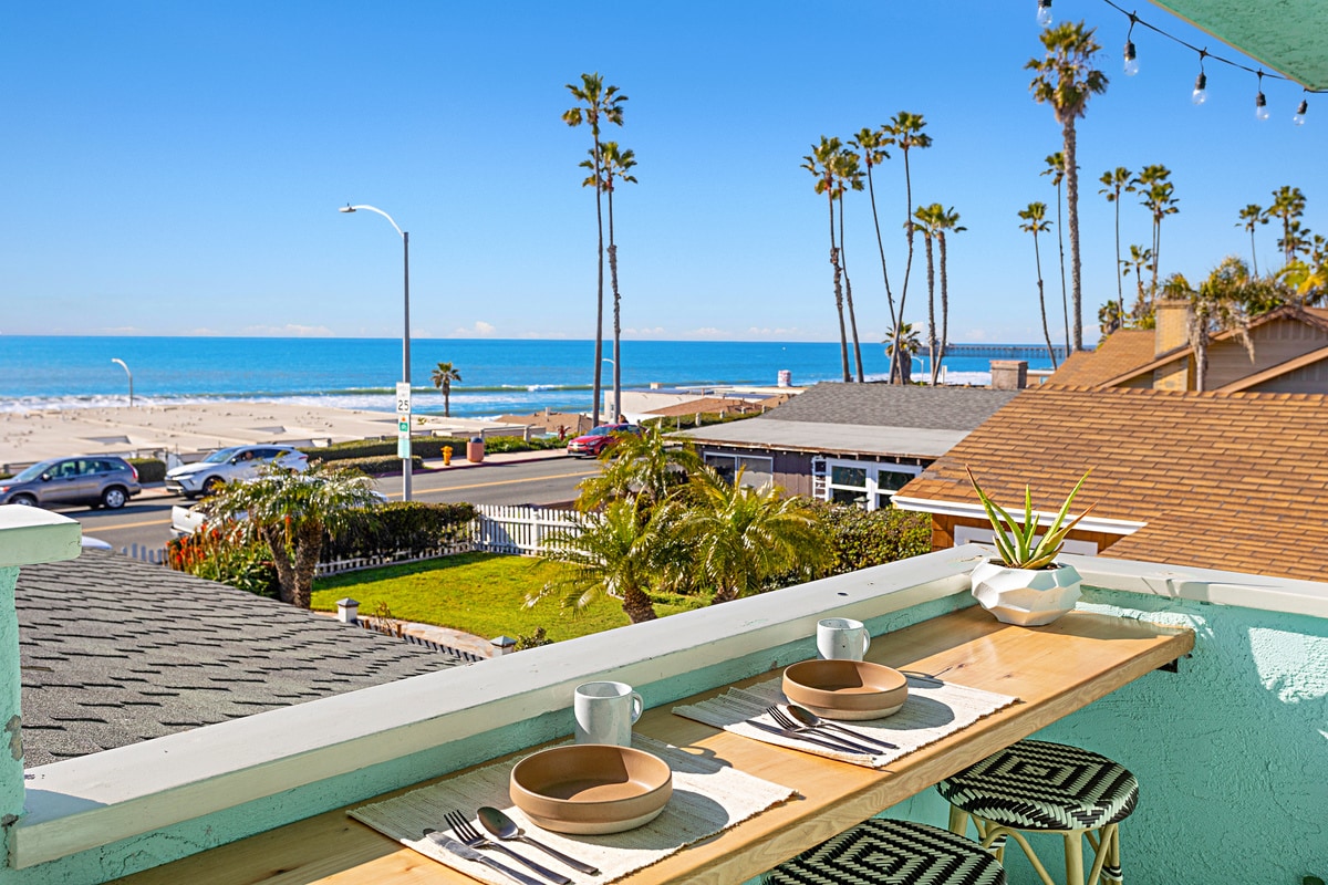 A semi-private front patio features a wooden table set for two, overlooking beautiful ocean views. Sandy beach and palm trees are visible in the background, with the calm waves gently lapping at the shore.