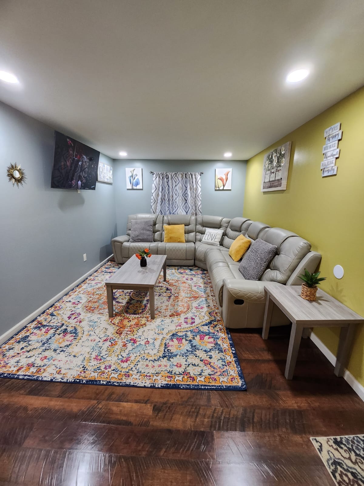 A comfortably arranged living area features a sizeable beige sectional sofa, a patterned area rug, and a coffee table. Art pieces adorn the walls, while a window is dressed with light curtains. Soft lighting is provided by recessed ceiling lights.