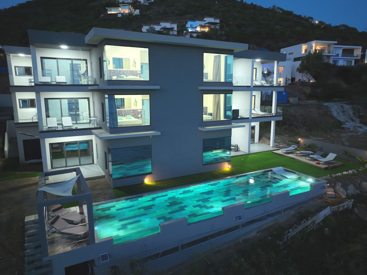 The modern villa is showcased from an aerial perspective, highlighting a long, illuminated swimming pool surrounded by greenery. Spacious balconies are seen on each floor, with contemporary outdoor seating. The building's sleek lines and large glass windows create a harmonious appearance in the evening light.