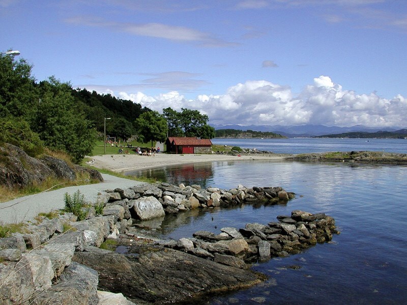 A serene coastal scene depicts a calm body of water reflecting clouds above. A sandy beach is framed by lush greenery and a small red structure stands nearby. Rocky formations lead into the water, creating natural patterns along the shore.
