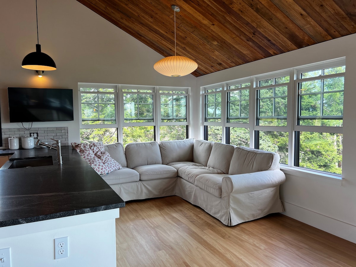 A spacious living area features a light-colored sectional sofa positioned near multiple windows, providing ample natural light. A modern pendant light hangs from the wooden ceiling, and a flat-screen TV is mounted on the wall. Views of greenery are visible through the windows.