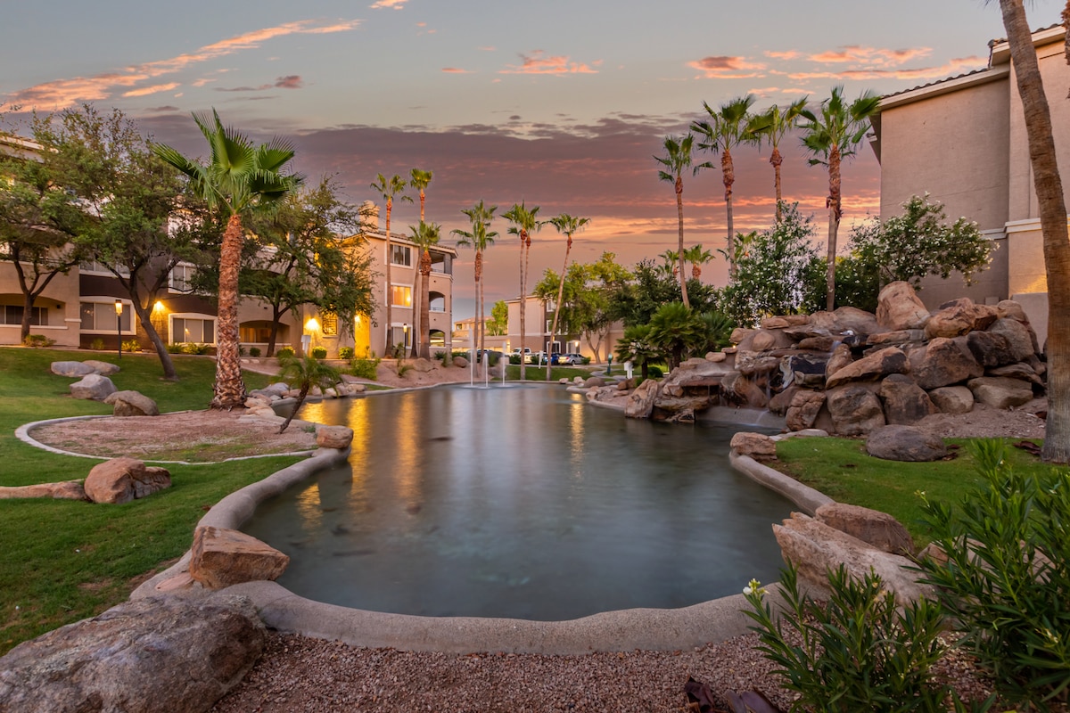 A serene lagoon-style pool is framed by lush greenery and palm trees, reflecting the warm tones of a sunset. The gentle flow of water over rocks creates a tranquil atmosphere, while the surrounding lights of the community buildings softly illuminate the scene.