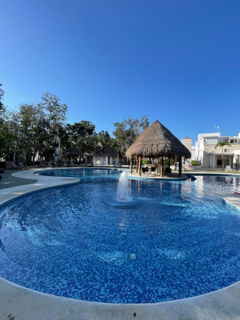 A spacious resort-style pool with a vibrant blue mosaic design features a thatched-roof palapa at its center. Surrounding the pool, lush greenery adds a natural touch to the inviting outdoor area, under a clear blue sky.