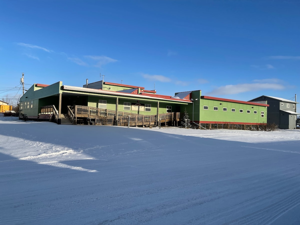 A spacious building is outlined against a clear blue sky, featuring a green exterior with red accents. Wooden decks line the front, leading to multiple entry points. Snow covers the ground, creating a wintery atmosphere.
