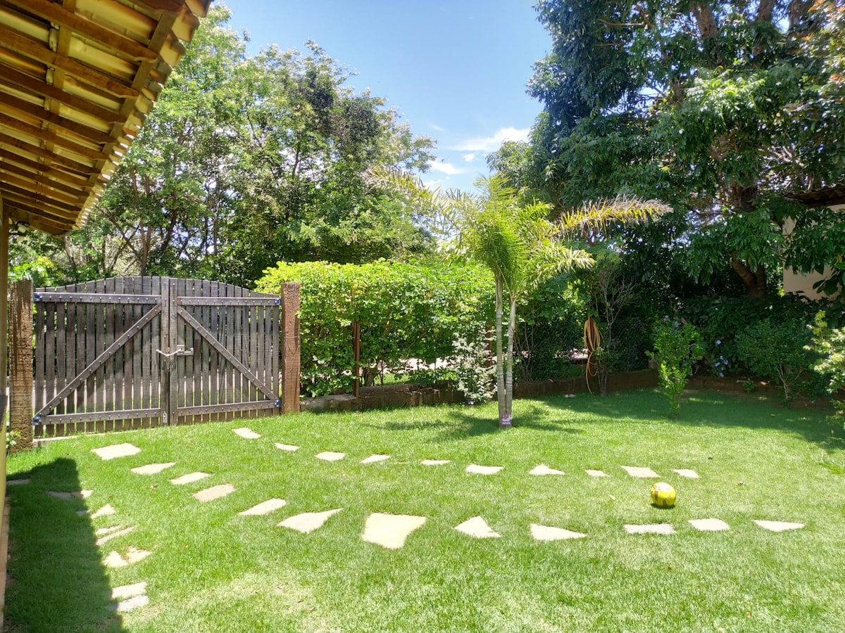 A spacious grassy area is framed by lush green bushes and trees. A stone pathway leads to a wooden gate, enhancing the openness of the space. Sunlight filters through the leaves, creating a bright and inviting atmosphere in the garden.