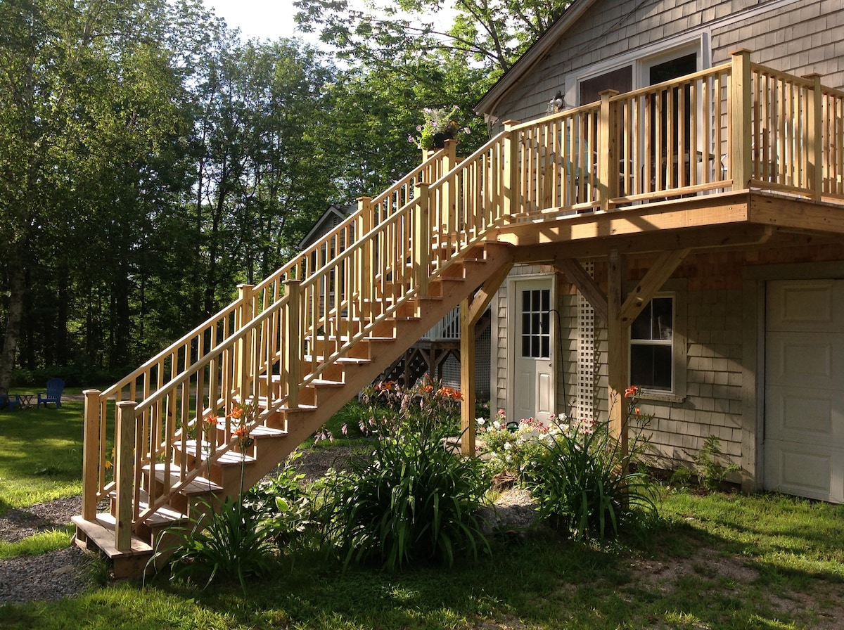 An exterior view showcases a wooden staircase leading to the cottage's second floor. The staircase is surrounded by a well-maintained garden with a variety of plants and flowers. Sunlight filters through the trees, highlighting the natural textures of the wood and greenery.