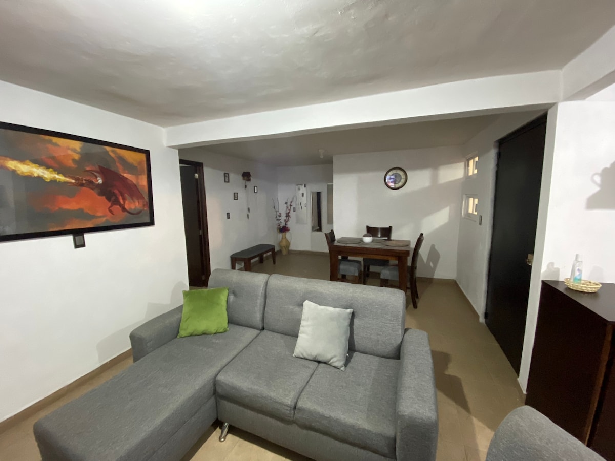An open living area is presented with a gray sectional sofa, complemented by a green accent pillow and a light gray cushion. A dining table for four is visible in the background, along with a decorative wall featuring artwork. The space is illuminated by soft lighting.