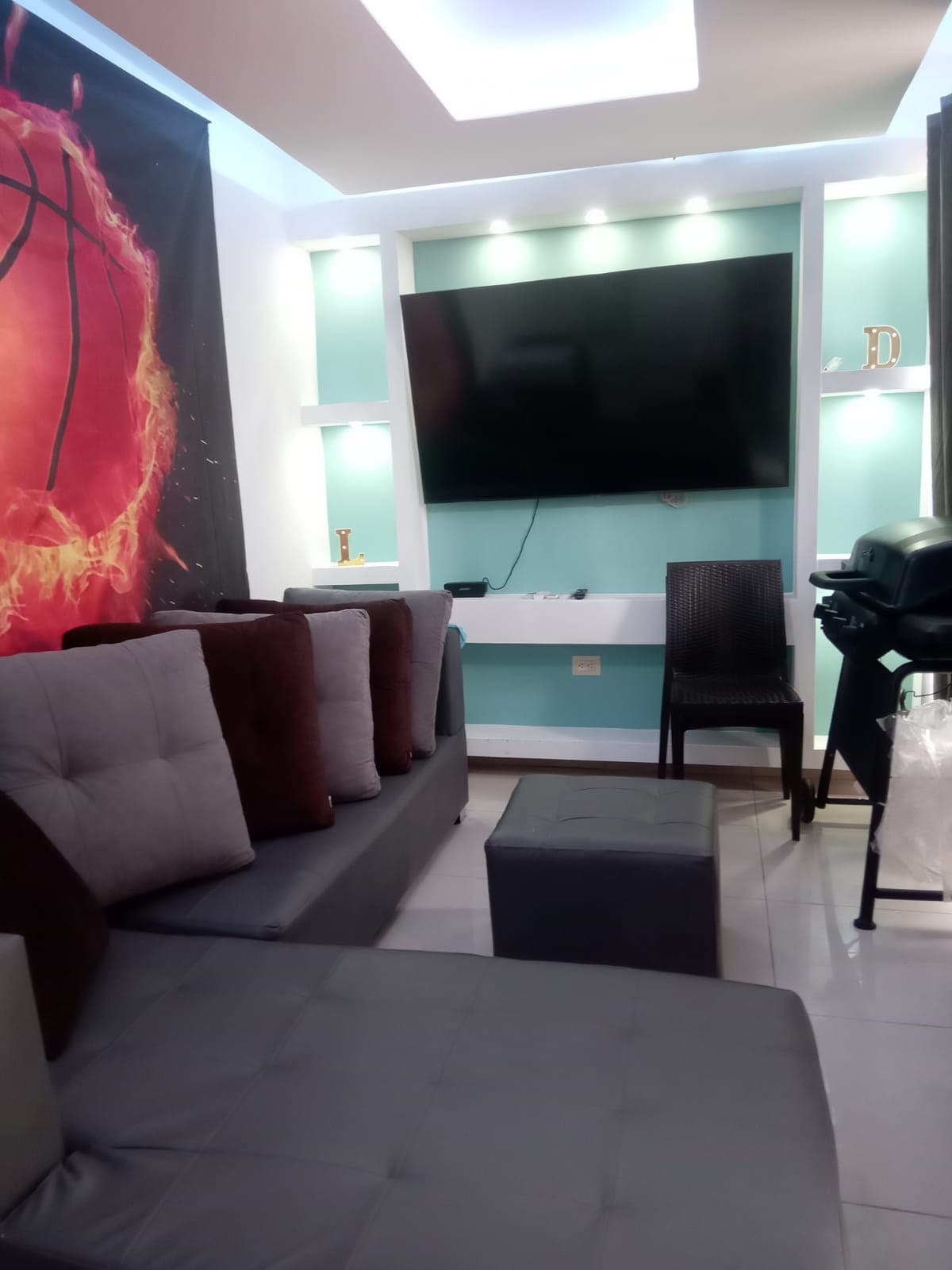 A modern living area features a comfortable sectional sofa with decorative cushions. A large wall-mounted television is positioned above a minimalistic media console. A small stool and a dining chair are included, with soft lighting highlighting the vibrant wall art in the background.