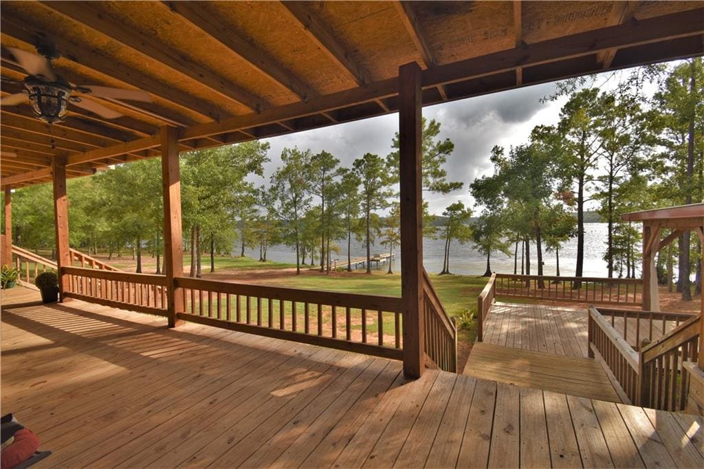 A spacious covered porch showcases wooden flooring and railings, providing views of the lake and surrounding trees. Steps lead down to the yard, revealing a green area that extends towards the tranquil waters. The scene is framed by tall pine trees, enhancing the outdoor ambiance.