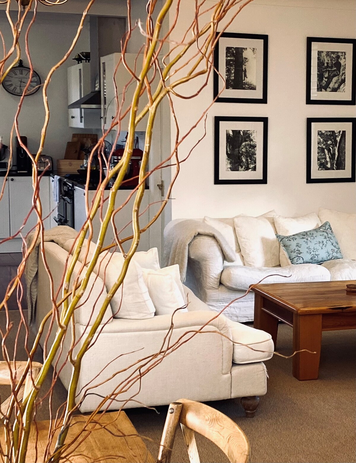 A cozy living area is showcased with cream-colored sofas adorned with cushions. A wooden coffee table sits in front, accompanied by a subtle arrangement of branches in a vase. Black-framed artwork is displayed on the walls, enhancing the room's charm.