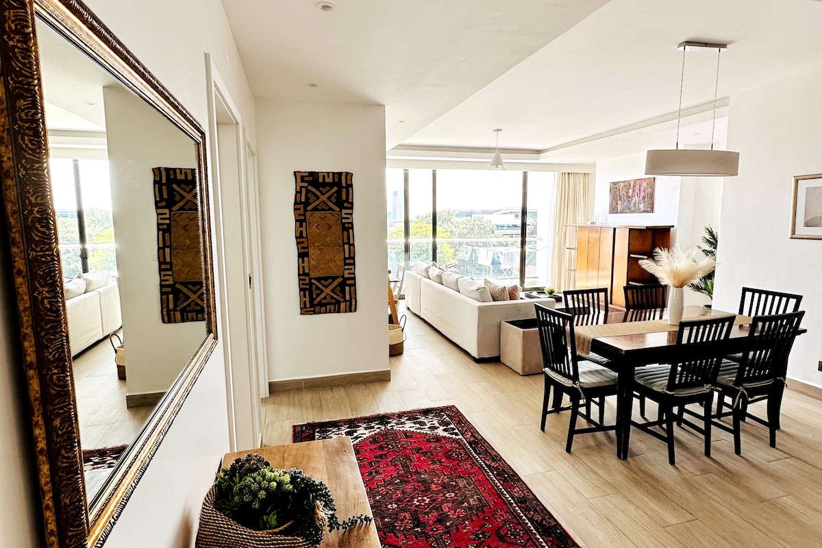 The open living and dining area showcases a dining table with black chairs set against a backdrop of large windows offering views. A stylish mirror and decorative wall art are positioned above a wooden table, enhancing the contemporary atmosphere. Natural light illuminates the space.