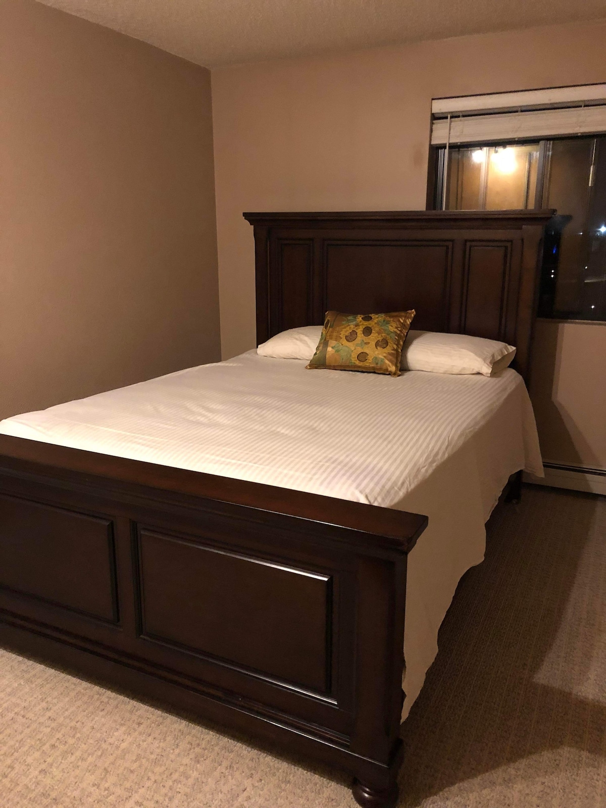 A spacious bed with a dark wooden frame is positioned against a light beige wall. An accent pillow with a floral design adds a pop of colour. Natural light filters in through a window, providing a subtle glow to the room.