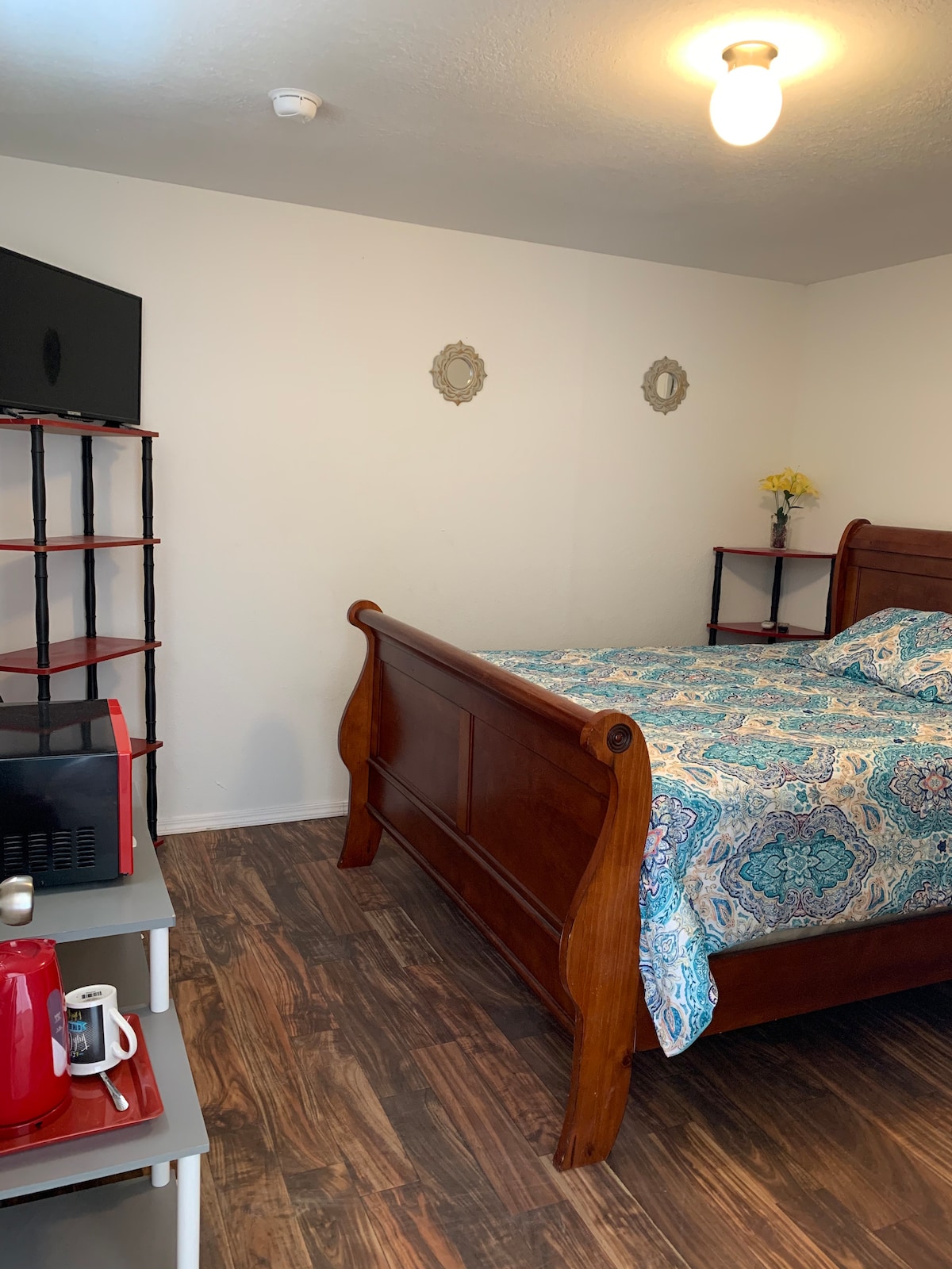 A bedroom features a wooden sleigh bed covered with a patterned comforter. A flat-screen TV is mounted on the wall across from the bed. A mini-fridge and microwave are situated on a shelf, with a small table displaying a kettle and cups nearby.