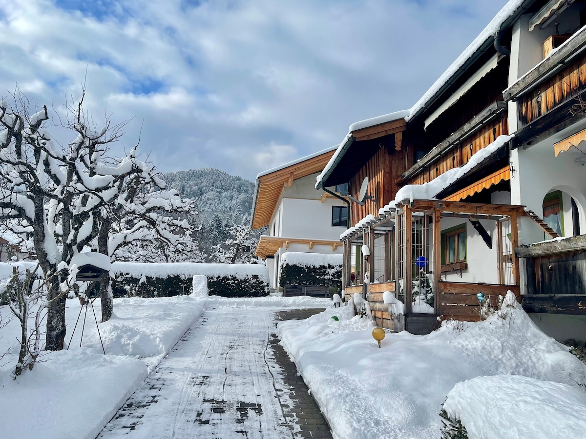 A snow-covered pathway leads to a charming building adorned with wooden details. The surrounding landscape features a tree bare of leaves and a backdrop of snow-clad mountains under a partly cloudy sky.