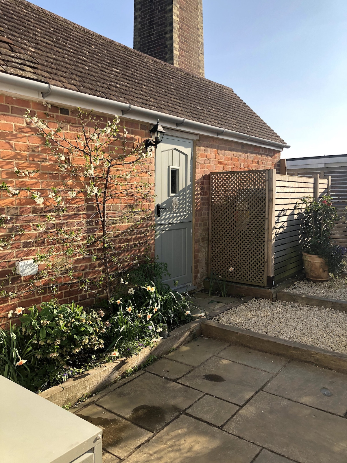 A charming outdoor space features a stone patio surrounded by greenery and flowering plants. A light-colored door is set within the rustic brick wall, and a wooden lattice panel adds visual interest. A small gravel area complements the inviting atmosphere of the courtyard.