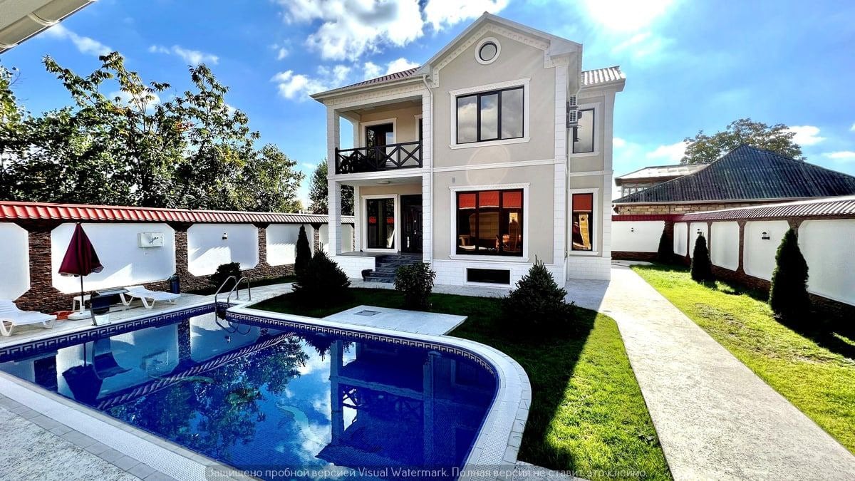 A spacious two-story house is surrounded by greenery, with large windows showcasing views of a refreshing pool. Lounge chairs and an umbrella are positioned near the pool, while a well-maintained pathway leads to the entrance, framed by neatly trimmed hedges.