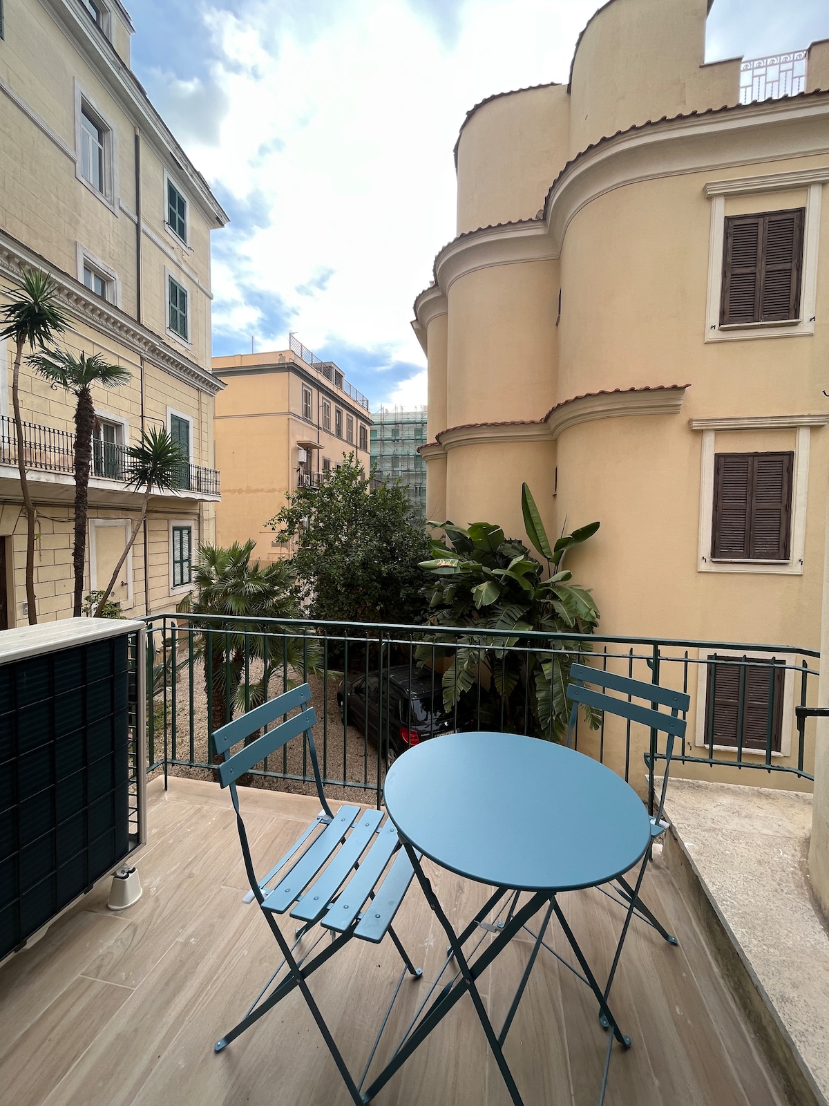 A small balcony features a round blue table accompanied by two folding chairs, offering a cozy outdoor seating area. Lush greenery and neighboring buildings are visible in the background, providing a sense of tranquility in a central location.