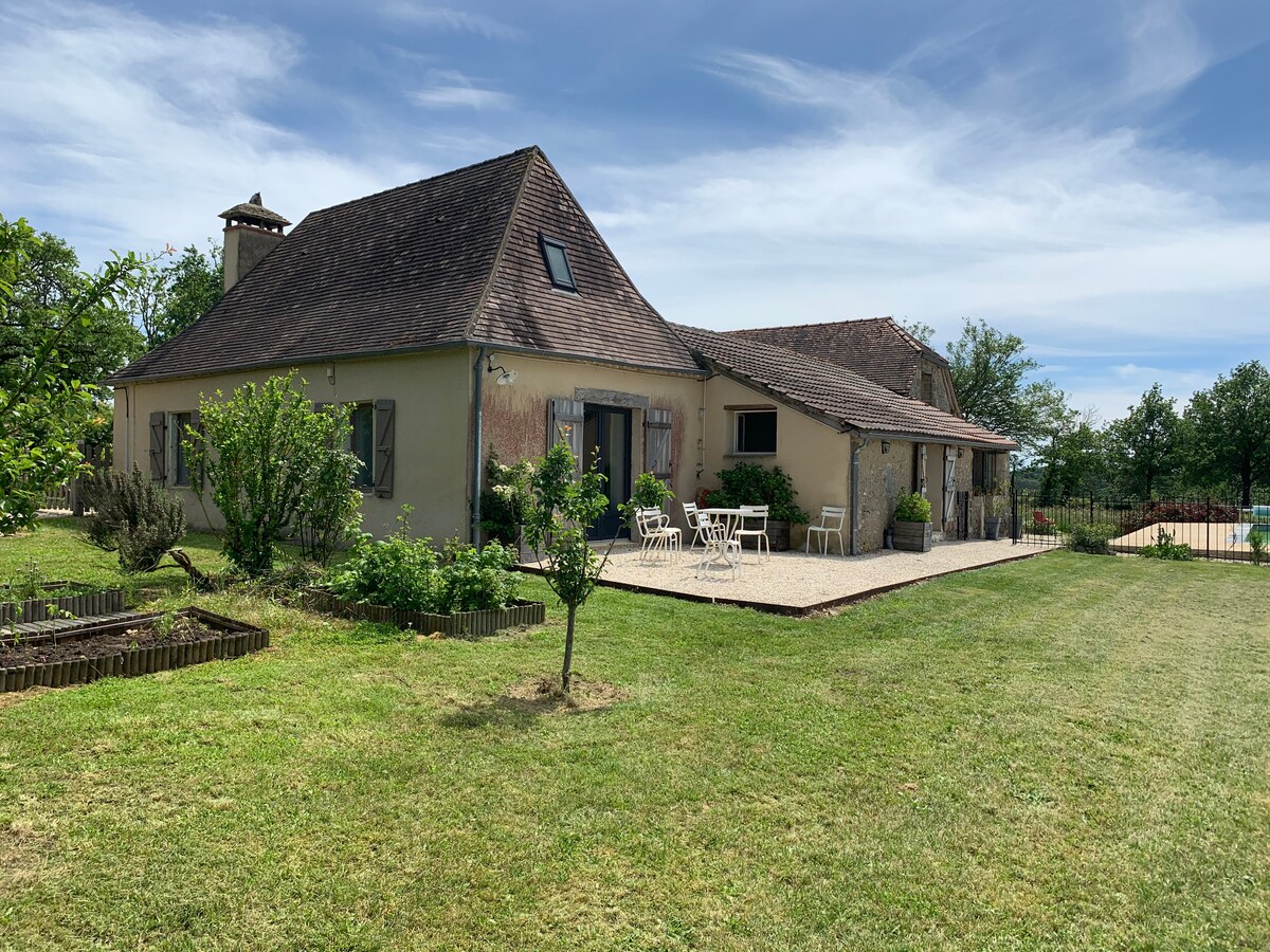The exterior view of a charming farmhouse features a sloped roof and neutral-colored walls. A seating area is set up on a stone-paved patio surrounded by greenery. The well-maintained lawn extends to a fenced area with a glimpse of the pool in the background.