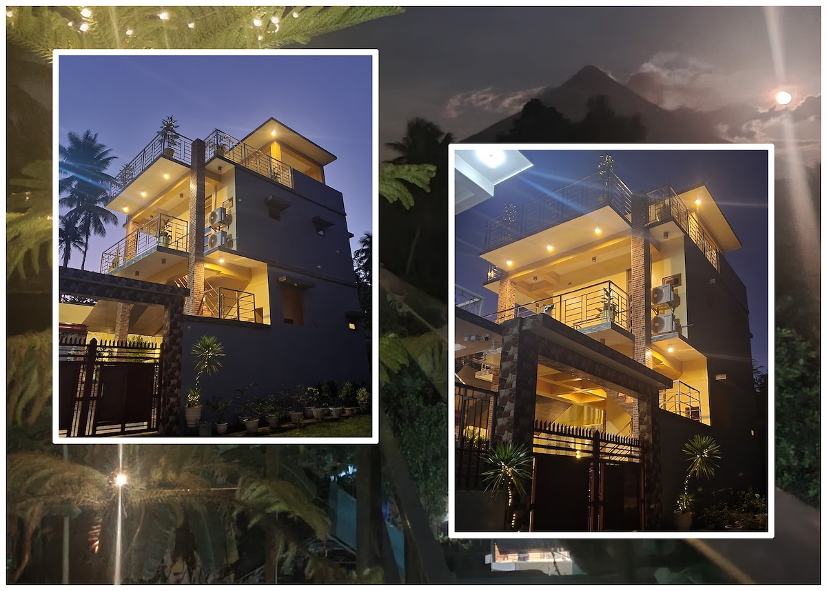 A contemporary three-story home is shown at twilight, featuring warm lighting that highlights its architectural details. The exterior is adorned with plants and a gated entrance, while balconies provide a view of the surroundings. The house merges modern design with a cozy ambiance.