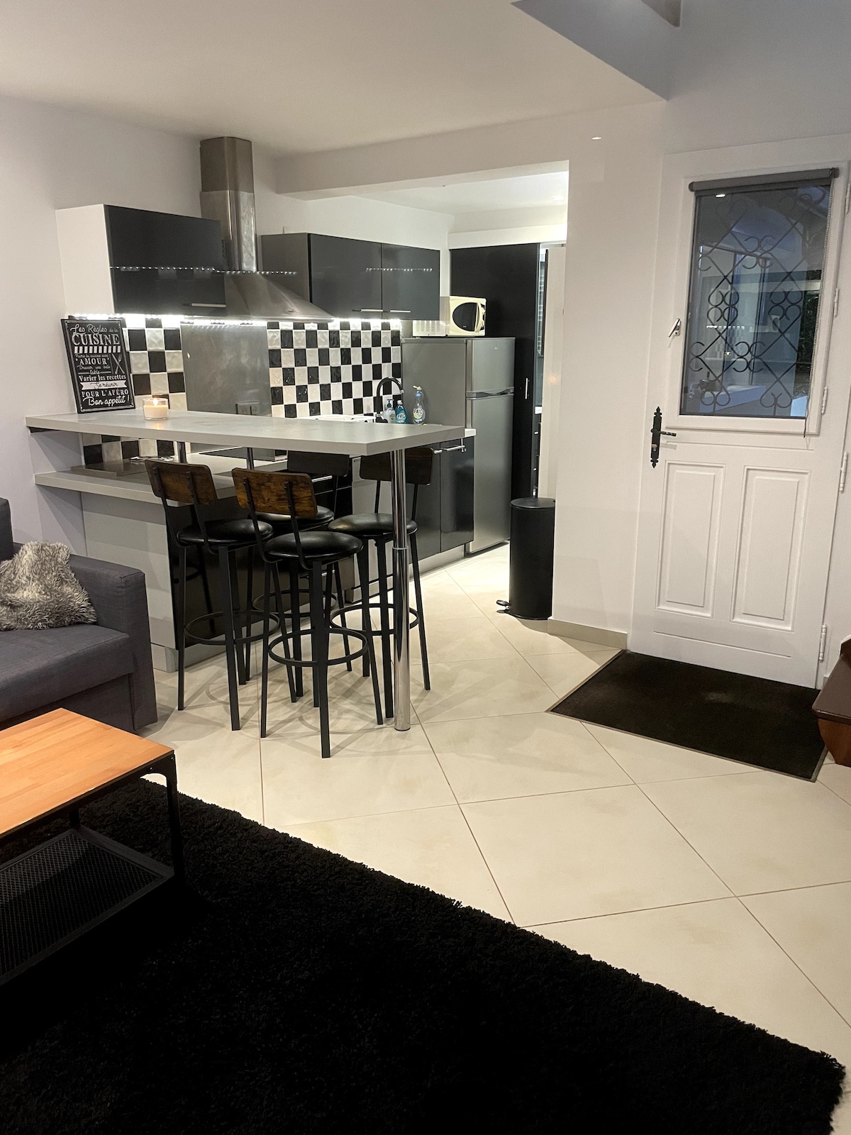 A modern kitchen area features a geometric black and white backsplash. The bar top is complemented by two black stools. An open layout displays a seating area with a grey sofa and a coffee table, adjacent to a door leading outside.