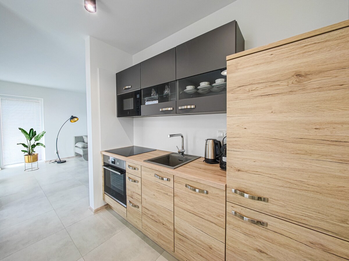 A modern kitchen space is featured, equipped with wood cabinetry and stylish dark countertops. Appliances include an oven, microwave, kettle, and refrigerator, providing essential functionality. Natural light brightens the area, complementing the open design, which connects to a living space visible in the background.