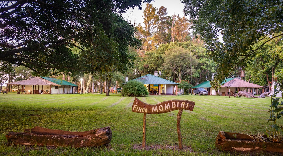 A sign with 'Finca Mombiri' is displayed prominently in a grassy area, surrounded by trees. Multiple cabins with green roofs are positioned around the landscape, showcasing a well-maintained outdoor space designed for relaxation and communal activities.