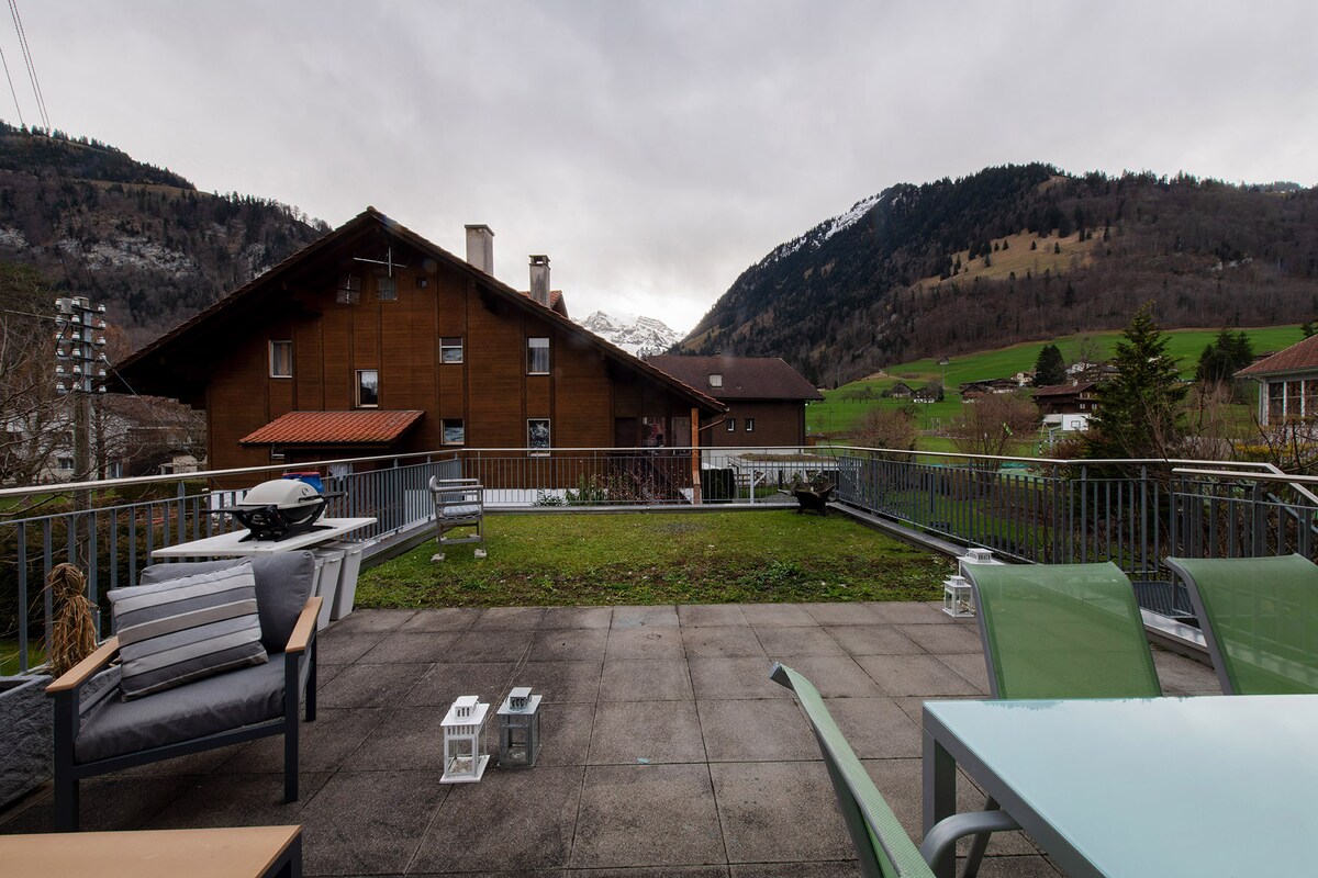 An outdoor terrace is presented, featuring a mix of seating arrangements, including lounge chairs and a dining table. The surrounding landscape showcases rolling hills and distant mountains, creating a serene backdrop. A gas grill and lanterns are visible on the patio, enhancing the outdoor space.