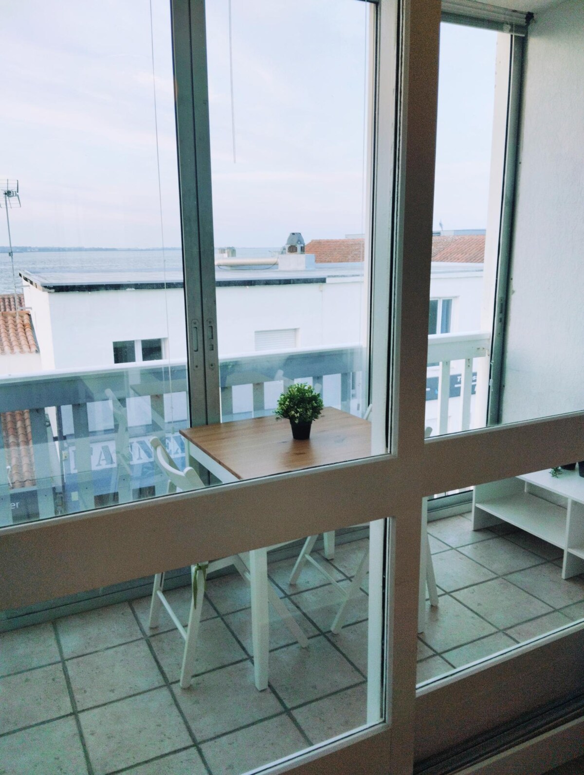 A bright balcony area is showcased, featuring a small wooden table and two white chairs. A potted plant sits center stage on the table. Large glass doors offer a view of the surrounding rooftops and the sea in the distance.