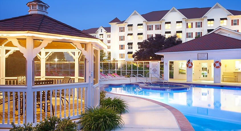 A well-maintained outdoor pool area features a gazebo with seating, surrounded by lush greenery. The pool is illuminated and offers a spa area, while the main building, showcasing multiple windows, stands in the background against an evening sky.