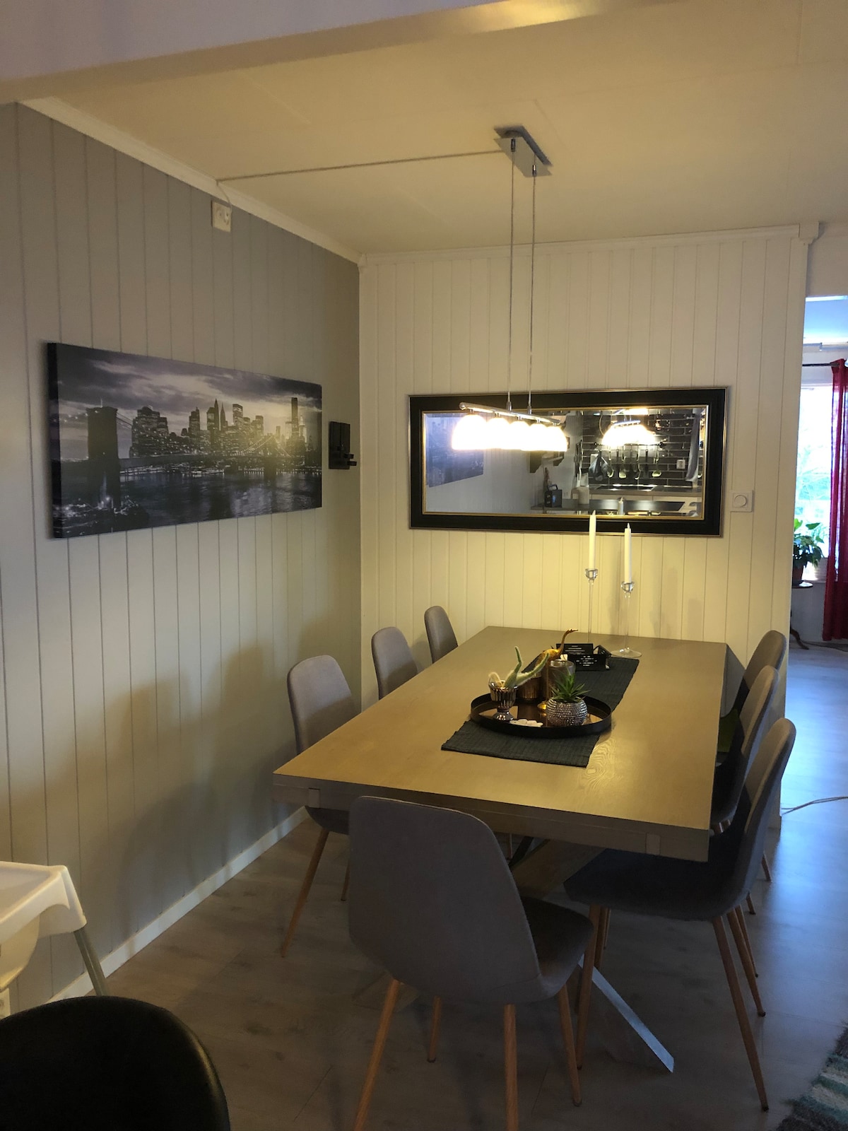 A dining area with a light wood table accommodates several chairs arranged around it. A black centerpiece is placed on the table. A large mirror reflects the room, and a photo of a cityscape is mounted on the wall, enhancing the space.