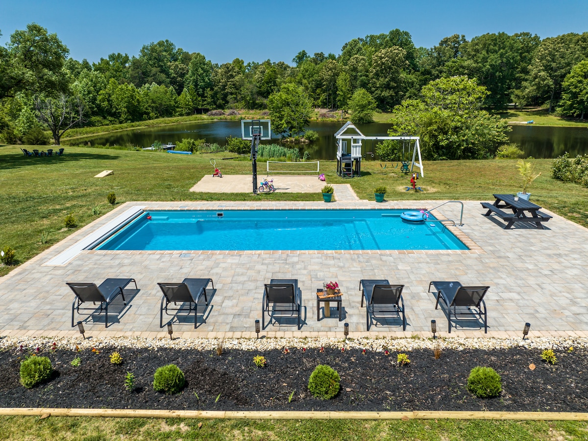 The swimming pool area features clear blue water surrounded by a spacious patio. Sun loungers are arranged neatly along one side, while a picnic table is positioned nearby. A playground and basketball hoop add to the outdoor recreation options, set against a backdrop of lush greenery.