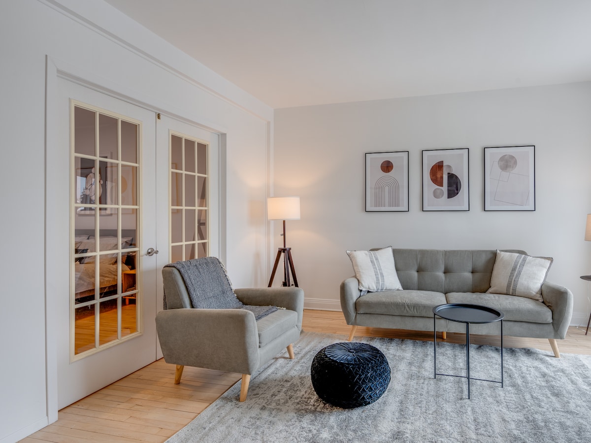 A bright living area features a soft grey sofa and a comfortable armchair, arranged around a small black coffee table. Light-colored walls are complemented by modern artwork, and a standing lamp adds gentle illumination. Double doors lead to another room, enhancing the open feel.