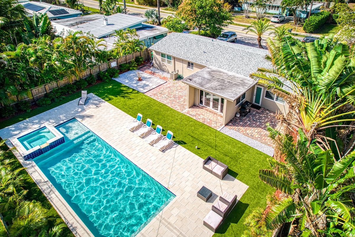 An aerial view of the property showcases a large, rectangular pool with a spa, surrounded by a tiled patio and vibrant green grass. Loungers are placed along one side of the pool, while lush palm trees provide a tropical feel to the outdoor space.