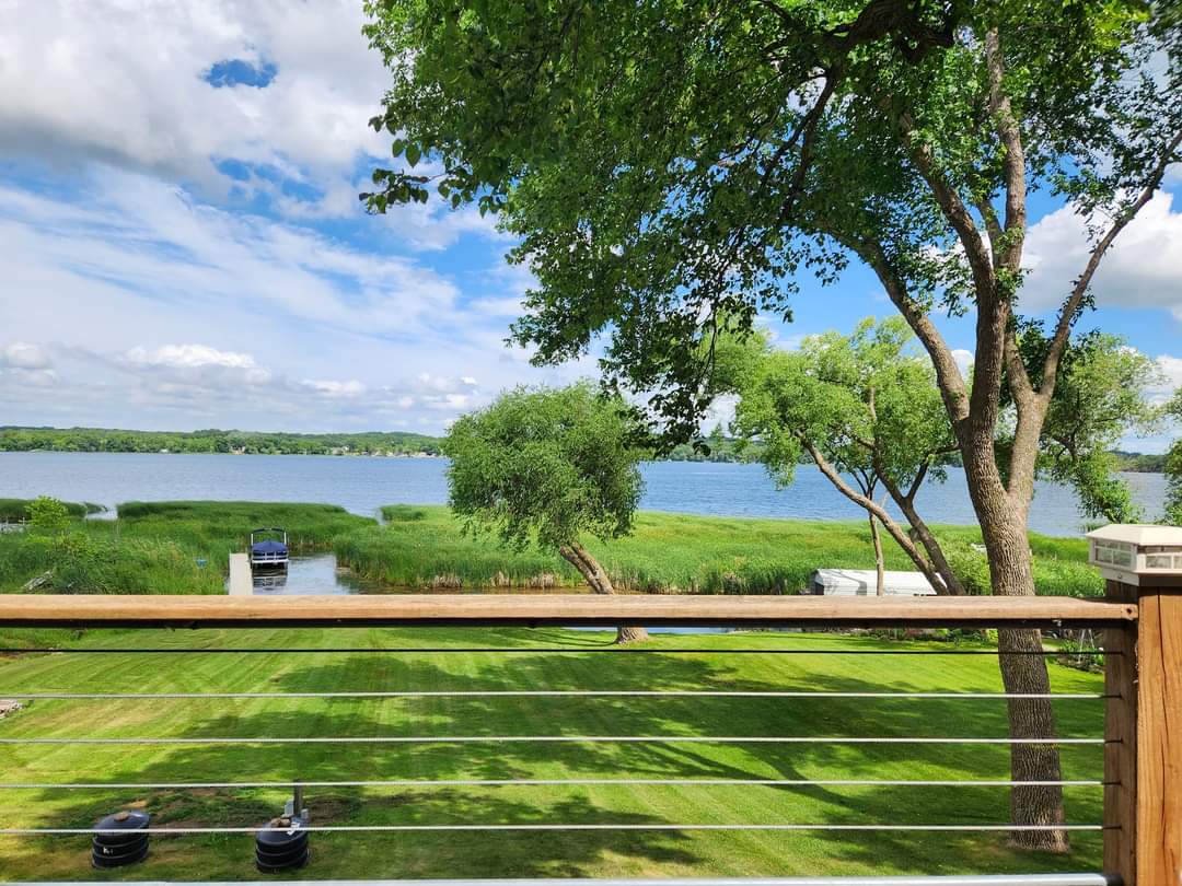 A view of Big Birch Lake is showcased from a wooden deck, framed by lush green trees and well-maintained grass. The calm water reflects the clouds and sky, while a boat is visible in the distance. The scene offers a peaceful lakeside atmosphere.