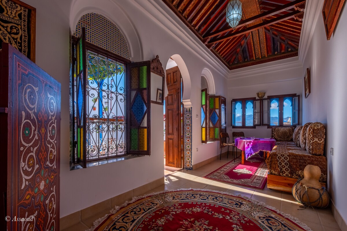 A warmly lit sitting area is adorned with intricate stained glass windows that provide colorful natural light. A traditional patterned rug covers the floor, while a wooden table is complemented by patterned cushions against the wall, creating a cozy and welcoming space.